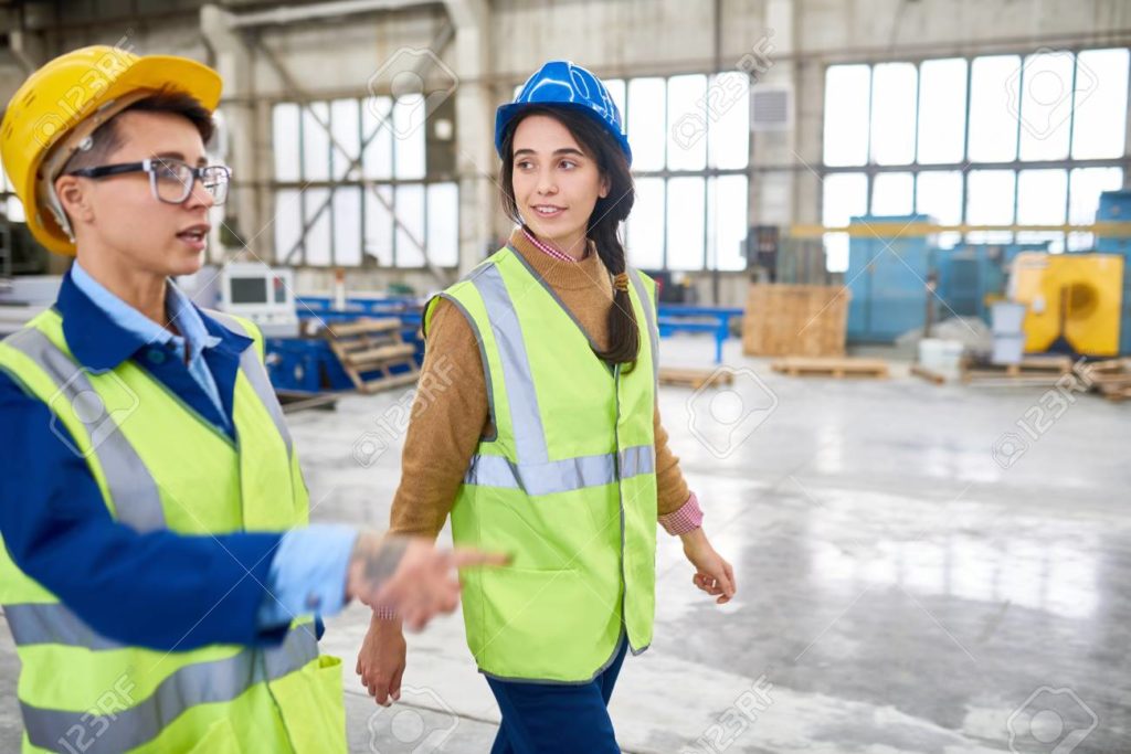 A non-binary person in a hardhat, walking beside a woman in a hardhat.