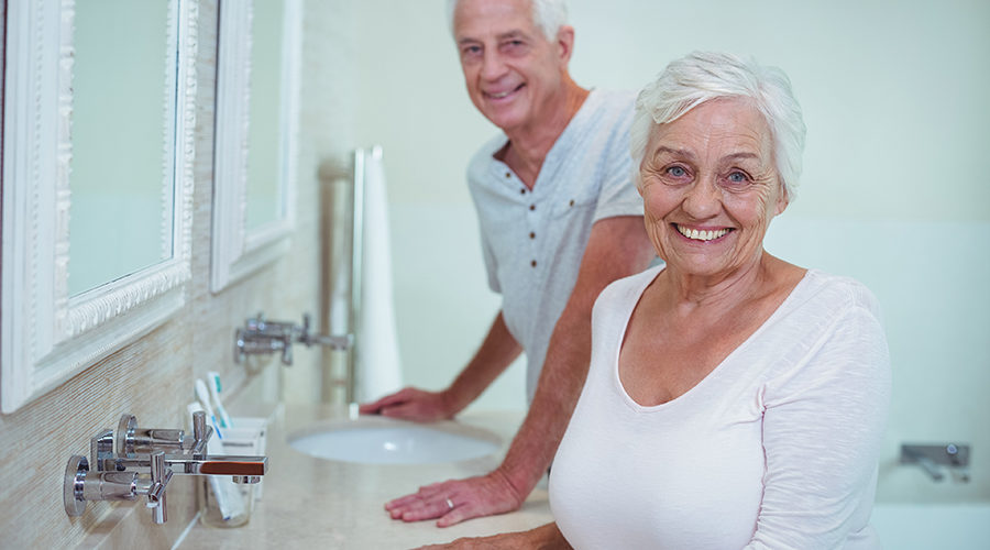 An elderly couple standing together at their bathroom sink, looking directly into the camera and smiling.