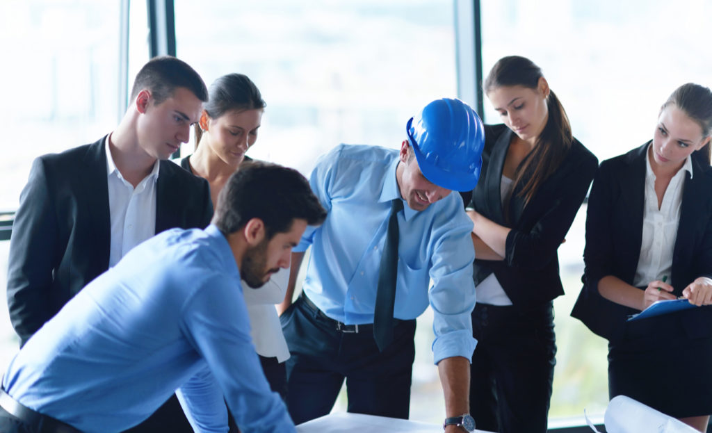 A team of professionals looking over blueprints, with one man in the center wearing a blue hard hat.